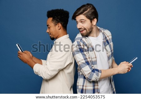 Side view young two friends happy men wear white casual shirts together hold in hand use mobile cell phone surfing internet peep isolated plain dark royal navy blue background People lifestyle concept