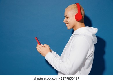 Side view young vivid fun dyed blond man of African American ethnicity wear white hoody headphones listen music use mobile cell phone isolated on plain dark royal navy blue background studio portrait
