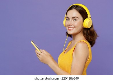 Side view young woman 20s wear yellow tank shirt headphones listen to music hold in hand use mobile cell phone look camera isolated on plain pastel light purple background. People lifestyle concept - Powered by Shutterstock