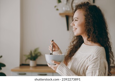 Side view young woman of African American ethnicity wear casual clothes sweater eat breakfast muesli cereals with milk fruit in bowl stand near table in kitchen at home. Lifestyle cooking food concept - Powered by Shutterstock