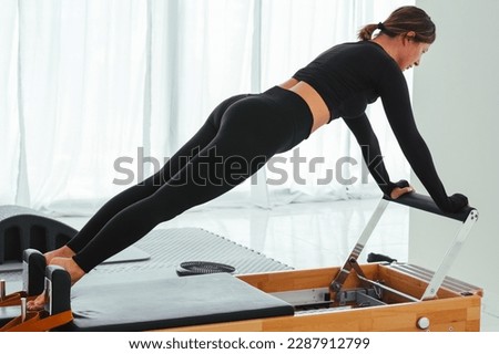 Side view of young woman in black sportswear doing plank position on pilates reformer machine in studio.