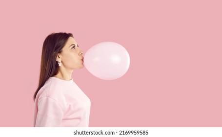 Side view of young woman blowing up pink balloon. Profile view of happy brunette girl inflating balloon on pink copy space studio background. Birthday party and fun concept