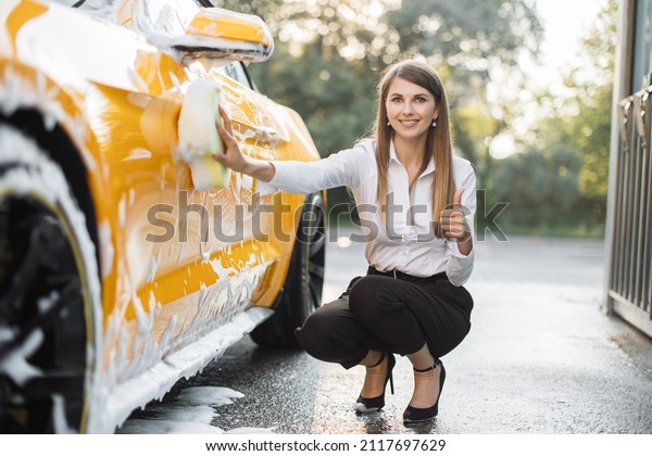 Side view of Young woman in business outfit,\
shirt and trousers using yellow sponge for cleaning car door with\
soap foam outdoors showing thumb\
up.