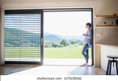 Side View Of Young Woman With Coffee Standing By Patio Door At Home.