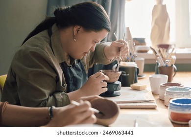 Side view of young woman with Down syndrome sitting at table in pottery workshop painting ceramic bowl, copy space - Powered by Shutterstock