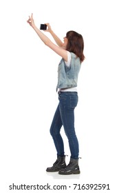 Side View Of Young Woman In Jeans Vest And Black Boots, Standing And Holding Cell Phone Over Her Head And Taking Photo. Full Length Studio Shot Isolated On White.