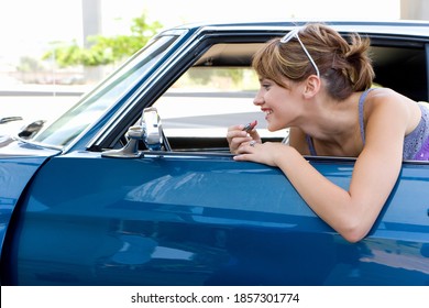 Side view of a young woman leaning on the door of a car applying lipstick in the rearview mirror. - Powered by Shutterstock