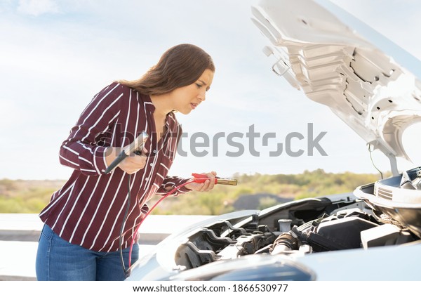 Side\
view of a young woman looking under her car hood to check the dead\
battery. Confused woman holding the jumper\
cables