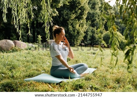Side view of young woman practicing yoga asana breathing exercise sitting in lotus position with closed eyes on sports mat in nature. Yoga and meditation, healthy active lifestyle.