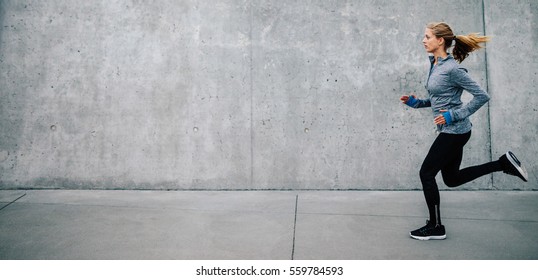 Side view of young woman running on sidewalk in morning. Health conscious concept with copy space.
