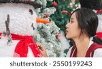 Side view of young woman staring eye-to-eye with orange-nosed snowman on background with Christmas tree and snow covered