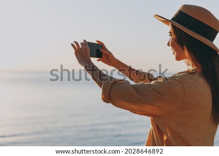 Side view young woman in straw hat shirt summer casual clothes take photo of landscape by mobile cell phone rest outdoors at sunrise sun dawn over sea beach. People vacation lifestyle journey concept