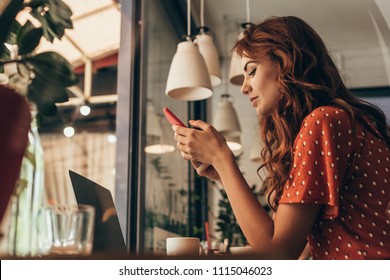 side view of young woman using smartphone at table with laptop in coffee shop - Powered by Shutterstock