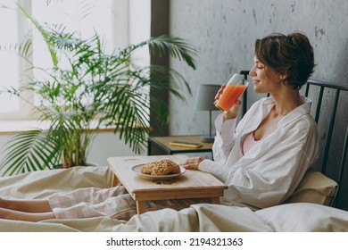 Side view young woman wear white shirt pajama she lying in bed eat breakfast drink orange juice rest relax spend time in bedroom lounge home in own room hotel wake up dream be lost in good mood day. - Powered by Shutterstock