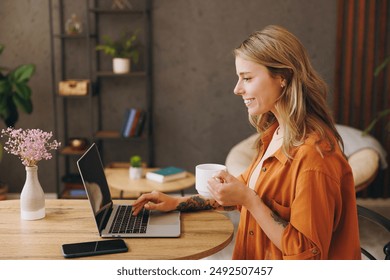 Side view young IT woman wearing orange shirt hold use laptop pc computer drink coffee sit alone at table in coffee shop cafe restaurant indoors work or study. Freelance mobile office business concept