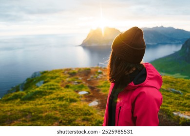 Side view of a young woman wearing a brown cap and red jacket, gazing at a stunning sunrise over mountains and a fjord. The serene landscape and soft light create a peaceful, breathtaking moment. - Powered by Shutterstock