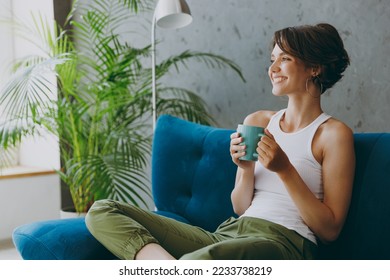 Side view young woman wears white tank shirt drink tea coffee sit on blue sofa couch stay at home hotel flat rest relax spend free spare time in living room indoors grey wall. People lounge concept - Shutterstock ID 2233738219