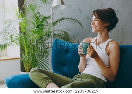 Side view young woman wears white tank shirt drink tea coffee sit on blue sofa couch stay at home hotel flat rest relax spend free spare time in living room indoors grey wall. People lounge concept