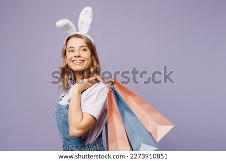 Side view young woman wears casual clothes bunny rabbit ears hold paper package bags after shopping isolated on plain light pastel purple background studio portrait. Happy Easter sale buy day concept