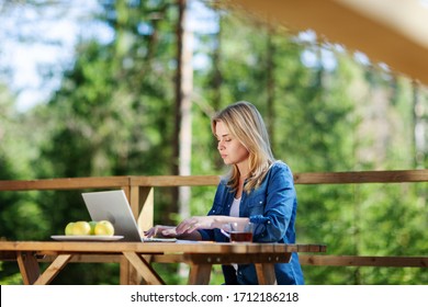 Side view of young woman working on laptop computer sitting at wooden table on balcony of countryside house - Shutterstock ID 1712186218