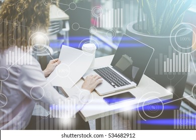 Side view,businesswoman sitting at table,on desk cup of coffee,laptop and smartphone,right hand woman clicks on touchscreen pc,in her left hand paper documents.On foreground digital charts,diagrams. - Shutterstock ID 534682912