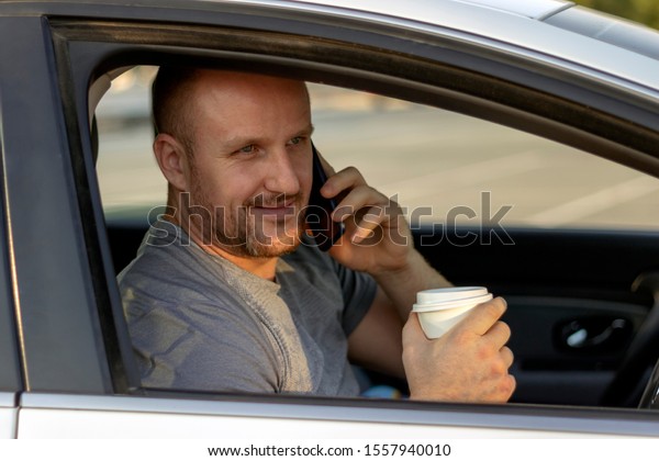 Side view,serious young man in the car while\
drinking coffee.Close up,sportsman in car using mobile.Handsome\
bearded male going to work in comfortable car.Man driving,drinking\
coffee and using phone.