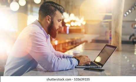 Side View.Young Bearded Businessman Wearing Blue Shirt,sitting At Table In Cafe And Uses Laptop.Nearby Is Smartphone And Cup Of Coffee. Man Checks Email On Computer. Freelancer Working Outside Office