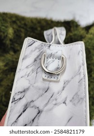 Side of white square shape bag with gray marble pattern with silver metal buckle accessory close up. Small size minimalist woman's handbag made of PU vegan leather. Green white background. Bag details
