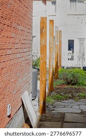 Side yard with brick wall, vertical wooden posts in a row, and a white house facade in background. Outdoor residential setting with walkway and garden area.