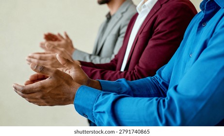 A side-angle shot of three men in formal wear, including a blue shirt, a maroon blazer, and a grey suit jacket, clapping their hands in unison.