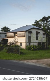 A side-angle view of a modest, elevated house in a lush tropical setting, featuring weathered siding and a classic silver corrugated metal roof. The structure is built on stilts with a set of wooden stairs leading to a side entrance, and it is bordered by a simple wire fence and verdant greenery. Bathed in the soft, low-angled light of late afternoon, the image captures a quiet, lived-in atmosphere characteristic of rural or subtropical residential architecture.