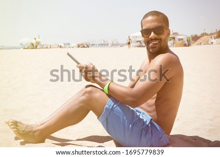 Sideshot of black man, feeling happy to spend his free time very well staying against blue sea and working with tablet.