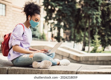 Side-View Of Black Student Girl Using Laptop Computer Wearing Earbuds And Protective Face Mask Having Distance Lesson Learning Online Sitting Outside. E-Learning, Virtual Lecture, Remote Study