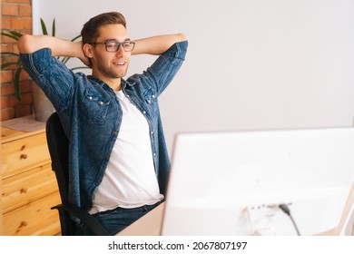 Sideview Of Cheerful Handsome Young Freelance Programmer Male In Stylish Glasses Holding Hands Behind Head Sitting At Desk With Desktop Computer At Home Office, Looking At Monitor Screen