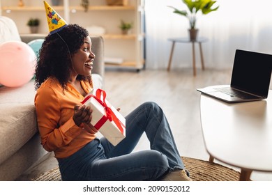 Side-View Of Excited African American Woman Having Online Birthday Party Talking To Laptop Video Calling, Holding Gift And Wearing Festive Hat At Home. Remote Holiday Celebration