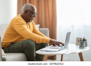 Side-View Of Mature Black Man Typing On Laptop Computer Browsing Internet Working Online Sitting On Couch At Home, Wearing Eyeglasses. Technology And Gadgets Concept