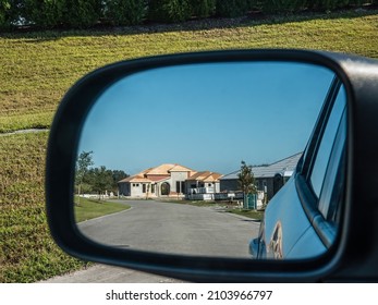 Side-view mirror on driver's side of car reflects perspective of suburban street with single-family houses under construction along one side on a sunny morning in southwest Florida