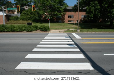 A side-view perspective of a pedestrian crosswalk, emphasizing the geometric pattern of the white stripes against the dark grey asphalt. The shot looks across the street towards a grassy verge and trees on the other side, with traffic lights visible in the distance. The low angle focuses on the texture of the road surface and the road markings, creating a leading line that draws the eye across the frame. Shadows from nearby objects fall across the foreground, adding depth and contrast to the scene. This image is a study in urban semiotics and safety infrastructure, illustrating the designated paths we create for movement. It covers themes of road safety, pedestrian rights, traffic laws, and the design of public spaces. The simplicity of the composition allows it to be used as a clean background for text or as a direct representation of city navigation and walkability.