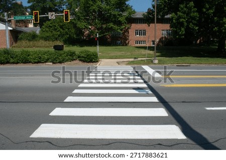 A side-view perspective of a pedestrian crosswalk, emphasizing the geometric pattern of the white stripes against the dark grey asphalt. The shot looks across the street towards a grassy verge and trees on the other side, with traffic lights visible in the distance. The low angle focuses on the texture of the road surface and the road markings, creating a leading line that draws the eye across the frame. Shadows from nearby objects fall across the foreground, adding depth and contrast to the scene. This image is a study in urban semiotics and safety infrastructure, illustrating the designated paths we create for movement. It covers themes of road safety, pedestrian rights, traffic laws, and the design of public spaces. The simplicity of the composition allows it to be used as a clean background for text or as a direct representation of city navigation and walkability.