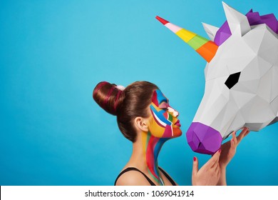 Sideview of pop art portrait of model wearing black opened top. Girl has saturated make up with bright geometrical figures and fancy hairdress. Posing on blue background with pink paper unicorn's head
