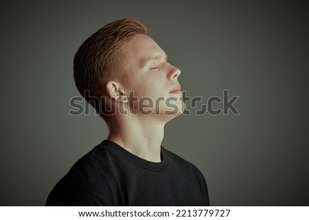 Sideview portrait of a handsome blond guy in a black t-shirt standing with closed eyes and taking a breath with a smile on his face. Grey background. Young people. 