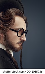 Sideview Portrait Of A Traditional Jewish Man With Sidedresses, In A Black Suit And Round Glasses. Studio Shot On A Dark Blue Background. 