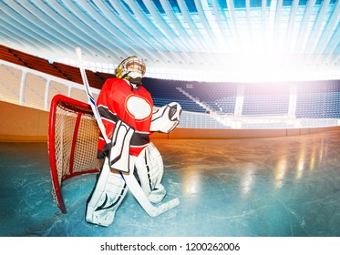 Side-view Portrait Of Young Goaltender Wearing Protective Equipment, Standing With Hockey Stick At Goal Crease On The Rink