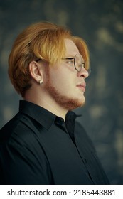 Sideview Portrait Of A Young Plump Man With Red Hair In A Black Shirt And Glasses Looking Pensively Away. Studio Shot. Modern Young Man.