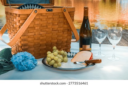 Sideview of a romantic setting in a dock next to a lake reflecting the tree colors with a picnic basket, a mockup friendly bottle of wine and two wine glasses, a plate with grapes and cheese and a touch of nature with a blue hydrangea on a white blanket. Background with copy space.