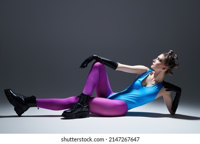 Sideview shot of a gorgeous model girl in bright extravagant clothes posing on the floor. Gray studio background. Black makeup. Modern vanguard fashion. 