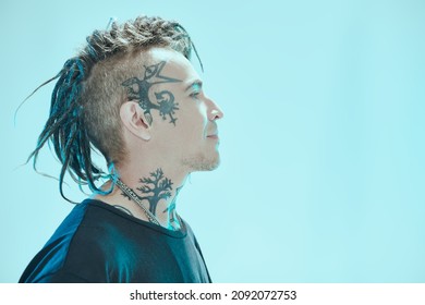 Sideview shot of a handsome  punk rock musician with dreadlocks and tattoos in black T-shirt smiling and looking away. Blue studio background. Youth alternative culture. Punk rock.