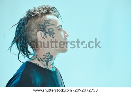Sideview shot of a handsome  punk rock musician with dreadlocks and tattoos in black T-shirt smiling and looking away. Blue studio background. Youth alternative culture. Punk rock.