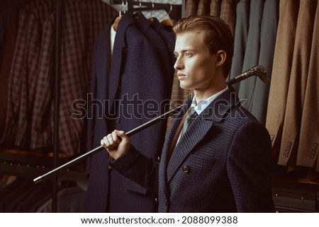 Sideview shot of a serious handsome blond man in elegant blue three-piece suit with a walking stick in his hand standing in the background of luxury classic suits in a store. Men's fashion and style.
