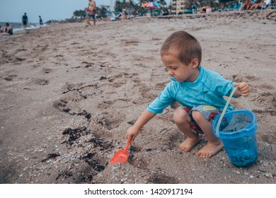 The Sideview Of A Small Baby Boy In A Blue Bathing Suit Sitting In The Sand And Shoveling Sand With His Red Shovel And Small Blue Bucket On The Beach 